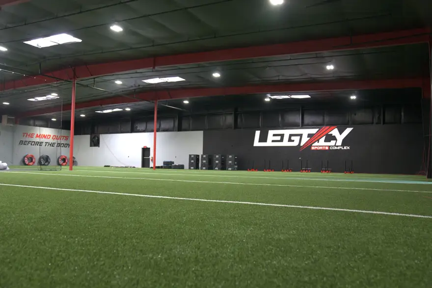 The indoor turf field at Milton Legacy, with the Legacy mark painted on the far wall and “The mind quits before the body” on the side wall.