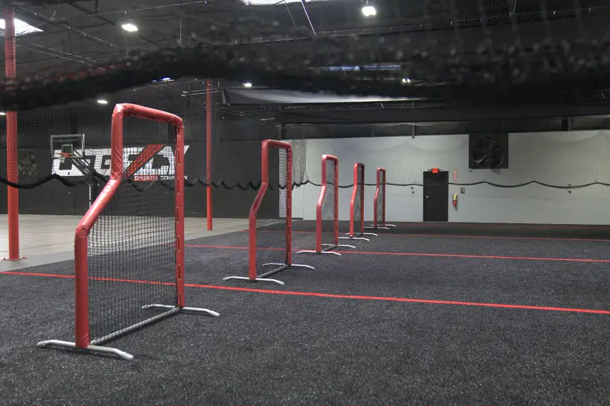 A netted training lane on the turf, divided by red-framed goals and hanging netting.